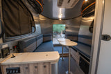 Modern RV rental interior with dinette, patterned benches, modular tables, and control panel. View outside through rear window.