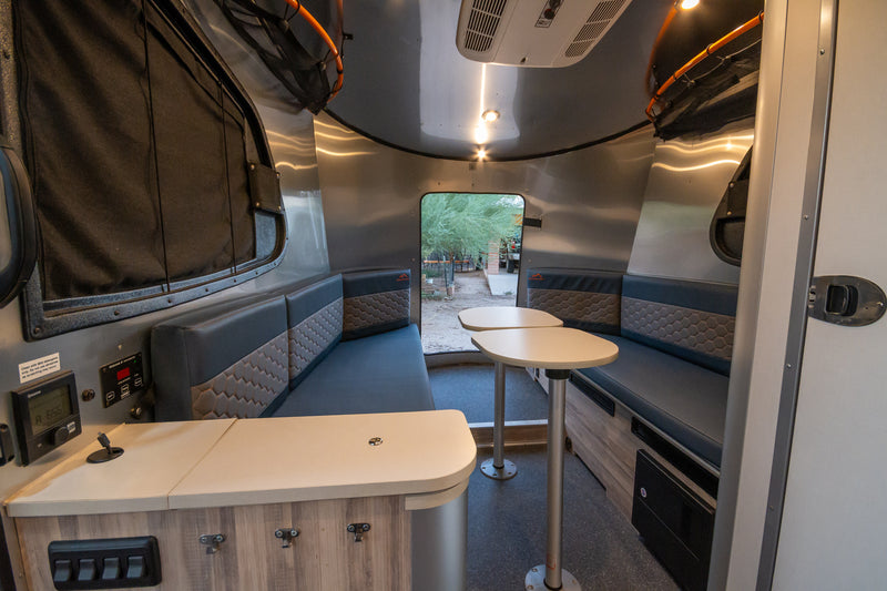 Modern RV rental interior with dinette, patterned benches, modular tables, and control panel. View outside through rear window. — photo 18