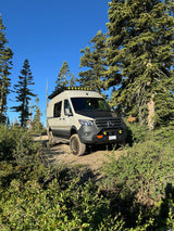 Explore off-grid! Light-colored off-road adventure van on a dirt trail among pines & bushes under a clear sky. Great for an RV rental.