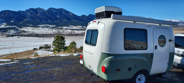 This white and green Happier Camper RV rental sits on a scenic overlook with snow-dusted mountains and valley in winter.
