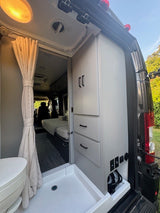 RV rental interior showcasing a compact bathroom with toilet, shower pan, storage cabinet, and a view into the living area with a bed.
