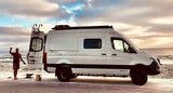 Enjoying the beach with an RV rental. A man stands beside a white Winnebago Revel camper van by the ocean at sunset.