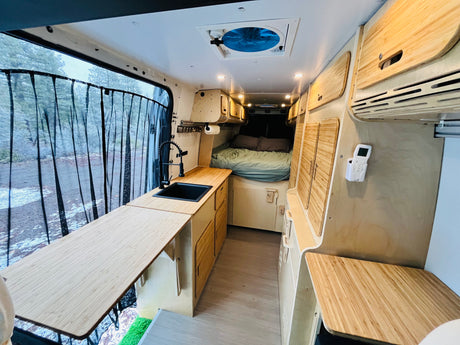 Modern rv rental camper van with light wood interior, kitchen, bed, and a scenic outdoor view.