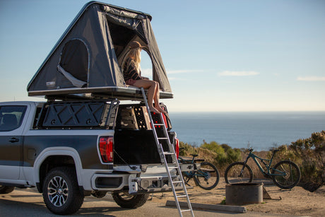 Scenic RV rental alternative: a pickup truck with a rooftop tent overlooking the ocean. A woman relaxes inside, bikes nearby for adventure.