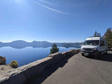 A white RV rental on a scenic road overlooking Crater Lake. Blue water reflects mountains under a clear sky.