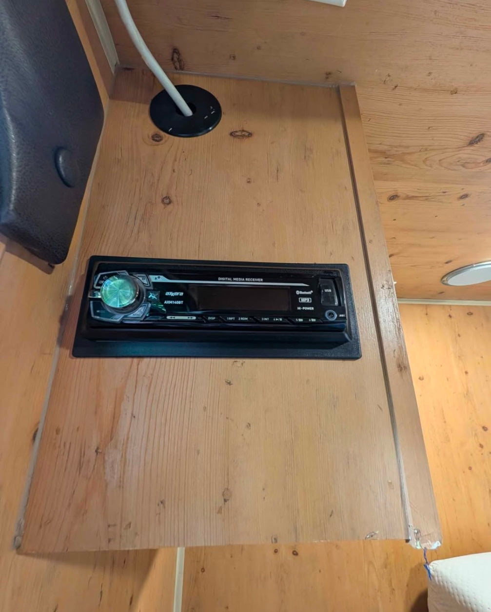 Inside an RV rental: a wooden wall with an Axerra car stereo installed. A white cable runs through a grommet above it.