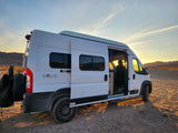 White Solis camper van for RV rental, open door reveals a traveler enjoying the desert sunset in a vast landscape.