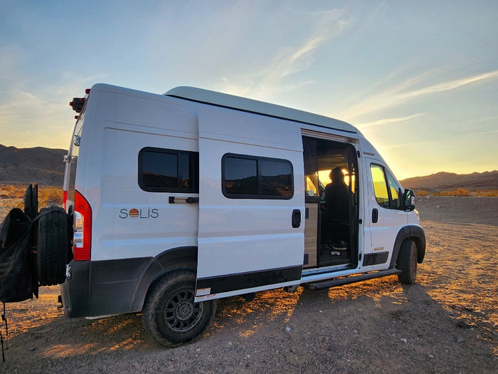 White Solis camper van for RV rental, open door reveals a traveler enjoying the desert sunset in a vast landscape.