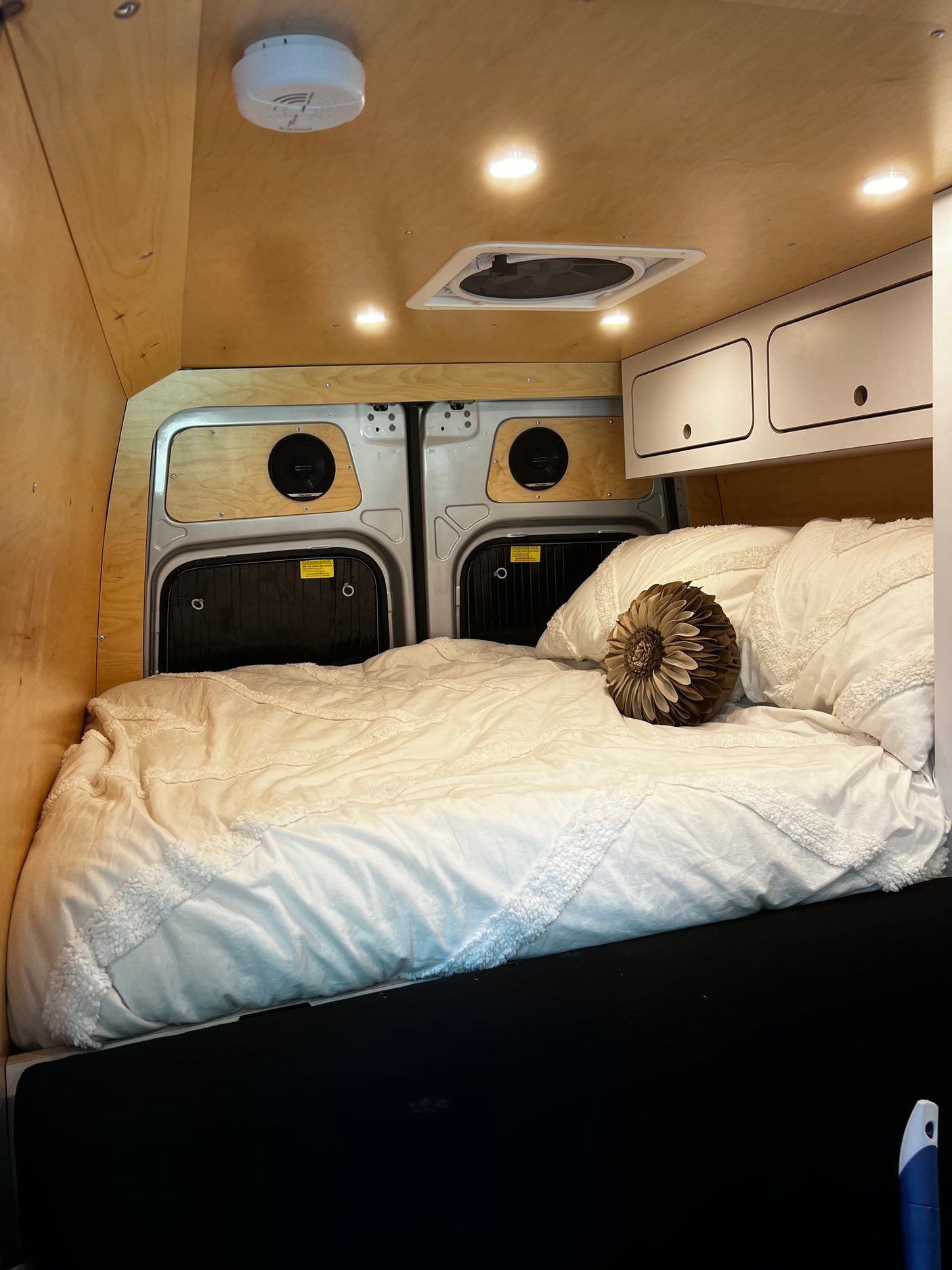 Inside a cozy RV rental: light wood walls, bright lights, ceiling fan, and a comfy bed with a decorative pillow. Perfect camper van setup.