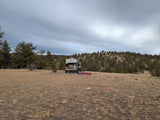 An RV rental travel trailer camped in a vast, dry field with pine-covered hills under a cloudy sky.