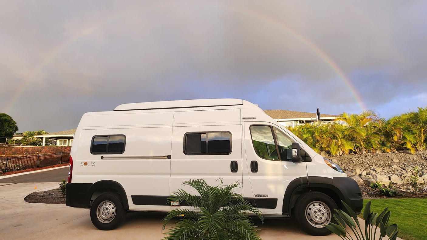 White Winnebago Solis RV rental parked outdoors, with a vibrant rainbow arching across the cloudy sky. Perfect for travel!