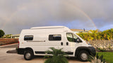 White Winnebago Solis RV rental parked outdoors, with a vibrant rainbow arching across the cloudy sky. Perfect for travel!