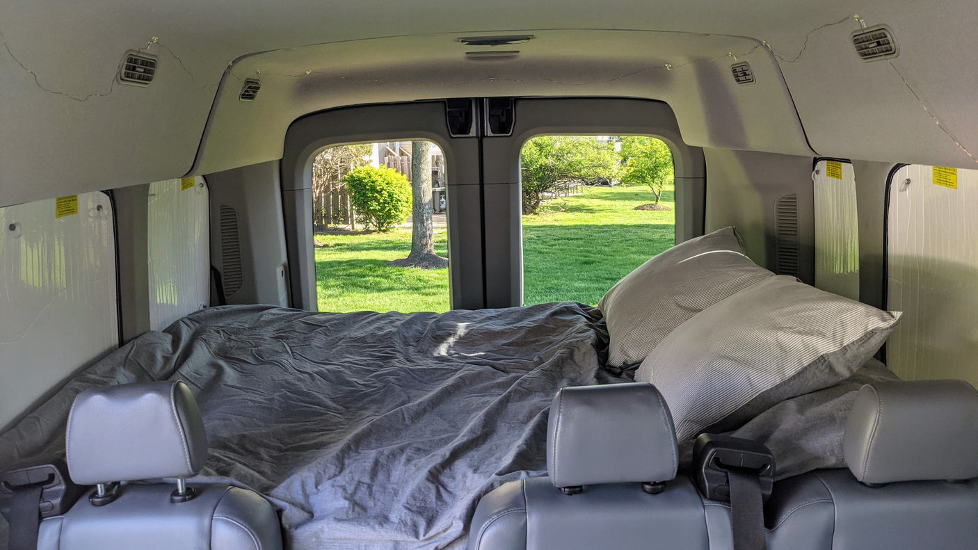 Inside an RV rental van: a comfy bed with pillows, seen from front seats. Green yard visible through the back doors.