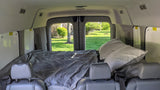 Inside an RV rental van: a comfy bed with pillows, seen from front seats. Green yard visible through the back doors.