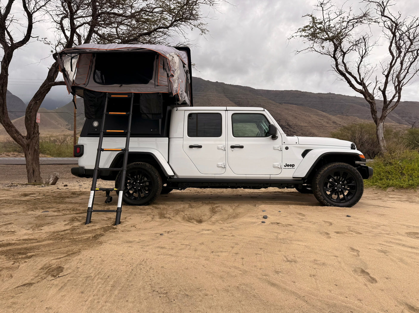 A white Jeep Gladiator with a rooftop tent, available for RV rental, parked on sand. Scenic outdoor camping setup.