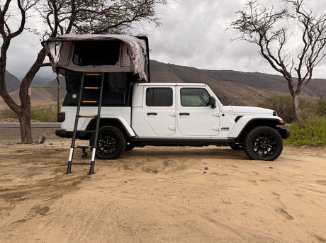 A white Jeep Gladiator with a rooftop tent, available for RV rental, parked on sand. Scenic outdoor camping setup.