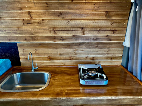 An rv rental's compact kitchen setup: wooden walls, counter, sink, and a portable stove for easy camp cooking.