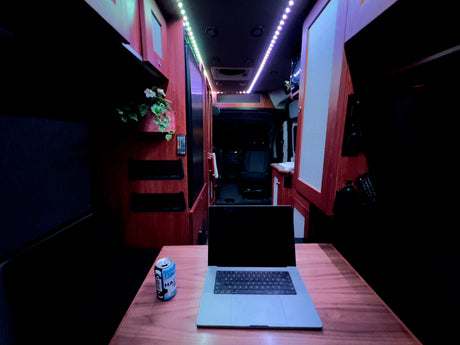 Inside an RV rental, a laptop & drink sit on a table. Colorful LED lights illuminate the cozy, dark interior, ideal for mobile work.