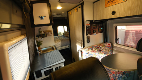 Inside a modern RV rental: a compact kitchen with sink, cozy bed beyond, and vibrant dinette seating. Control panel visible.