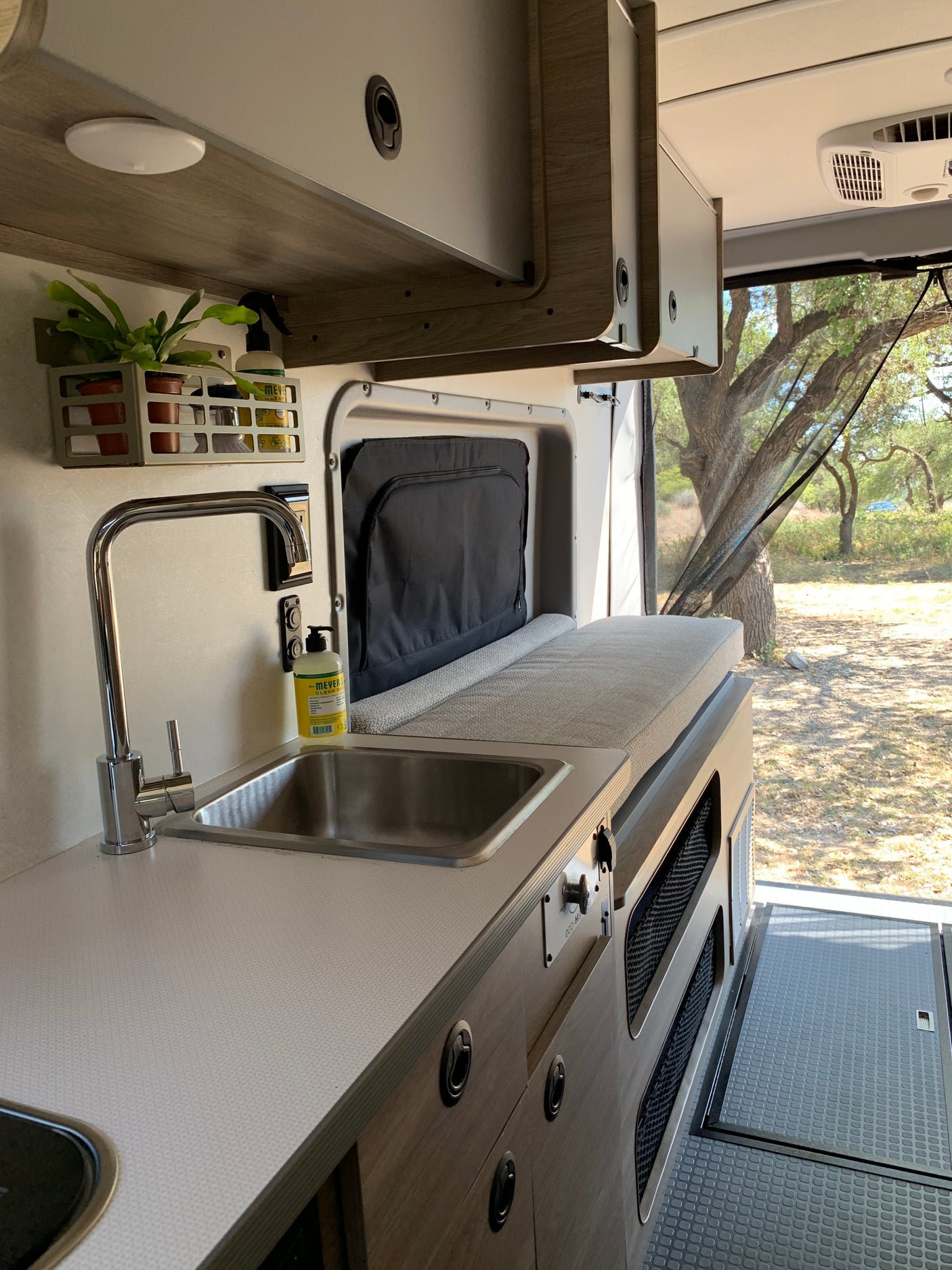 Modern RV rental interior with kitchen sink, overhead storage, comfy bed, and a large window overlooking nature.