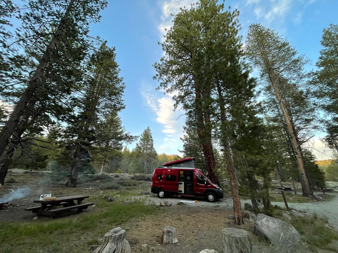Red RV rental parked in a tranquil forest campsite with tall pine trees, a picnic table, and campfire smoke rising.