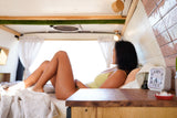 Woman relaxes on bed inside modern RV rental. Bright interior, wood & white accents, ideal for van life.