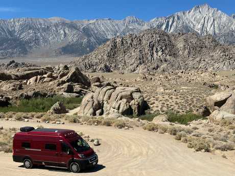 Red RV rental parked on a winding dirt road, nestled among rocky hills with towering gray mountains in the background.