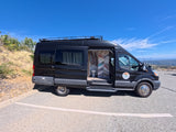 Black RV rental camper van with open side door revealing interior toilet & colorful wall, parked with mountain views.