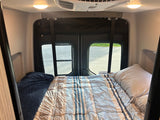 An RV rental's cozy camper van interior. A bed with striped comforter, rear windows, overhead lights & AC for comfortable travel.