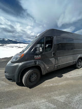 Rugged Ram ProMaster rv rental on a dirt road, amidst snowy mountains under a cloudy sky, ready for adventure.