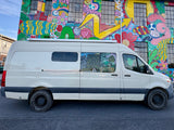 Brightly painted building mural behind a white camper van (RV rental) with unique window drawings. Parked on asphalt.