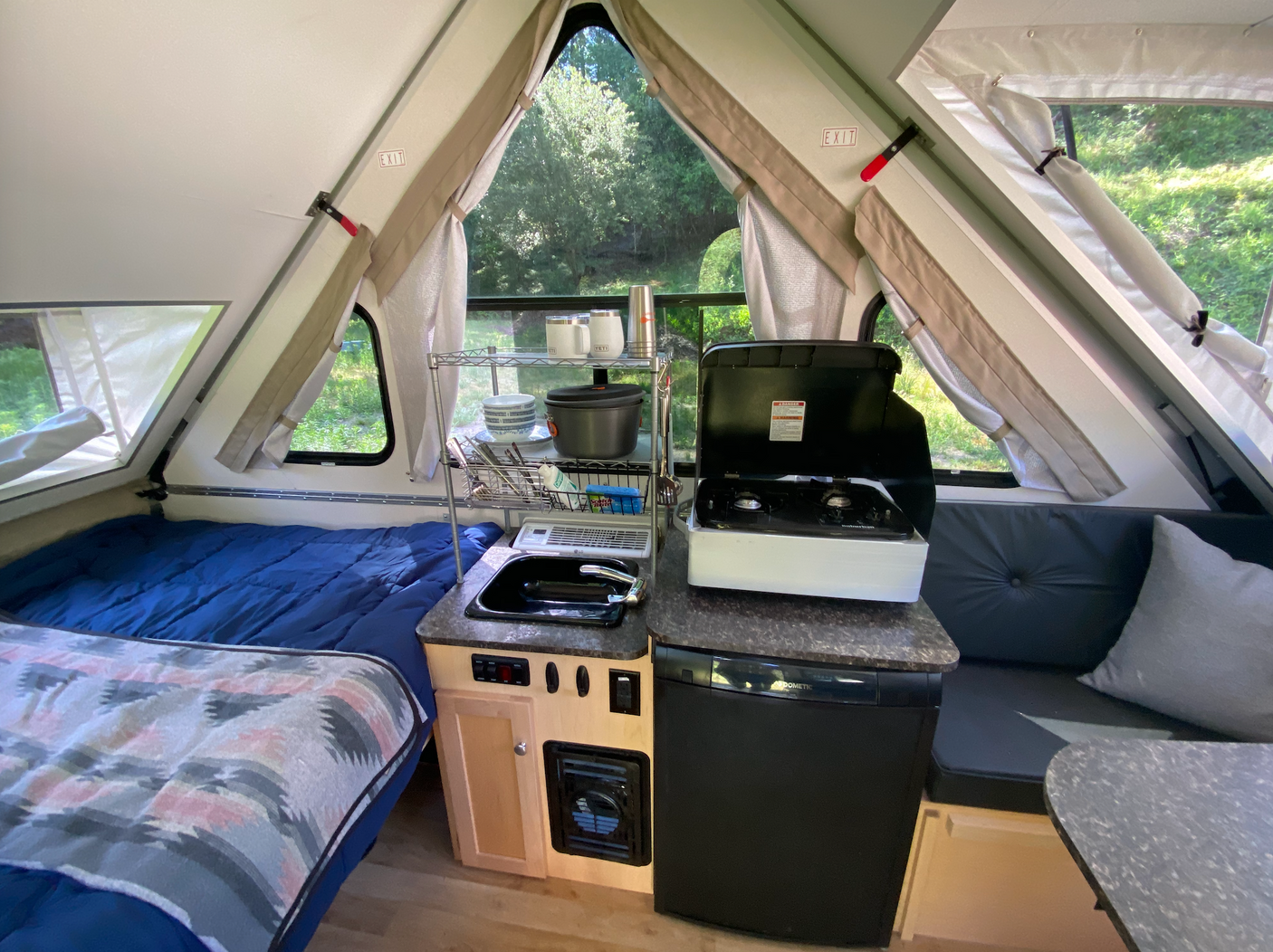 Cozy A-frame rv rental interior showcasing a bed, kitchenette with sink & stovetop, and seating area, surrounded by nature views.