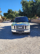 Front view of a green Ford van, excellent for RV rental, parked on gravel under a sunny blue sky.