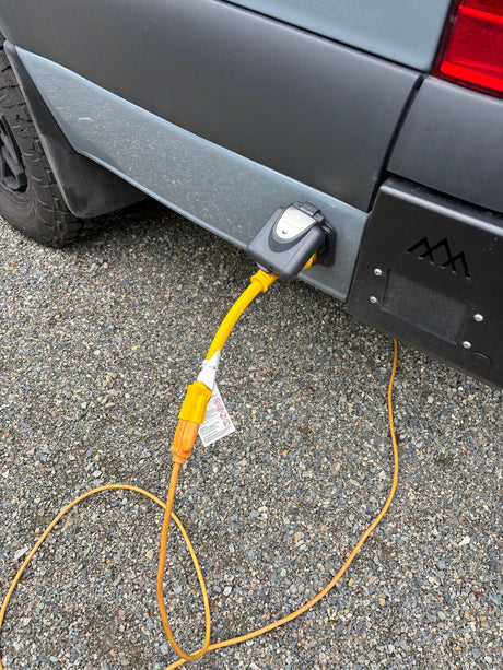 Grey RV rental van plugged into external power on gravel via yellow extension cord.