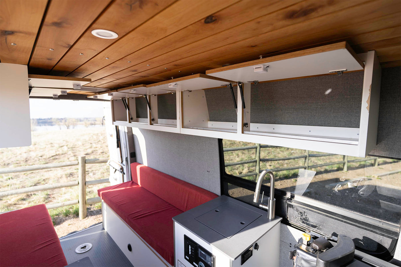 Interior of an RV rental with wooden ceiling, open white overhead cabinets, red bench seating, and a compact kitchen sink.