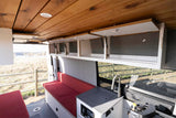 Interior of an RV rental with wooden ceiling, open white overhead cabinets, red bench seating, and a compact kitchen sink.