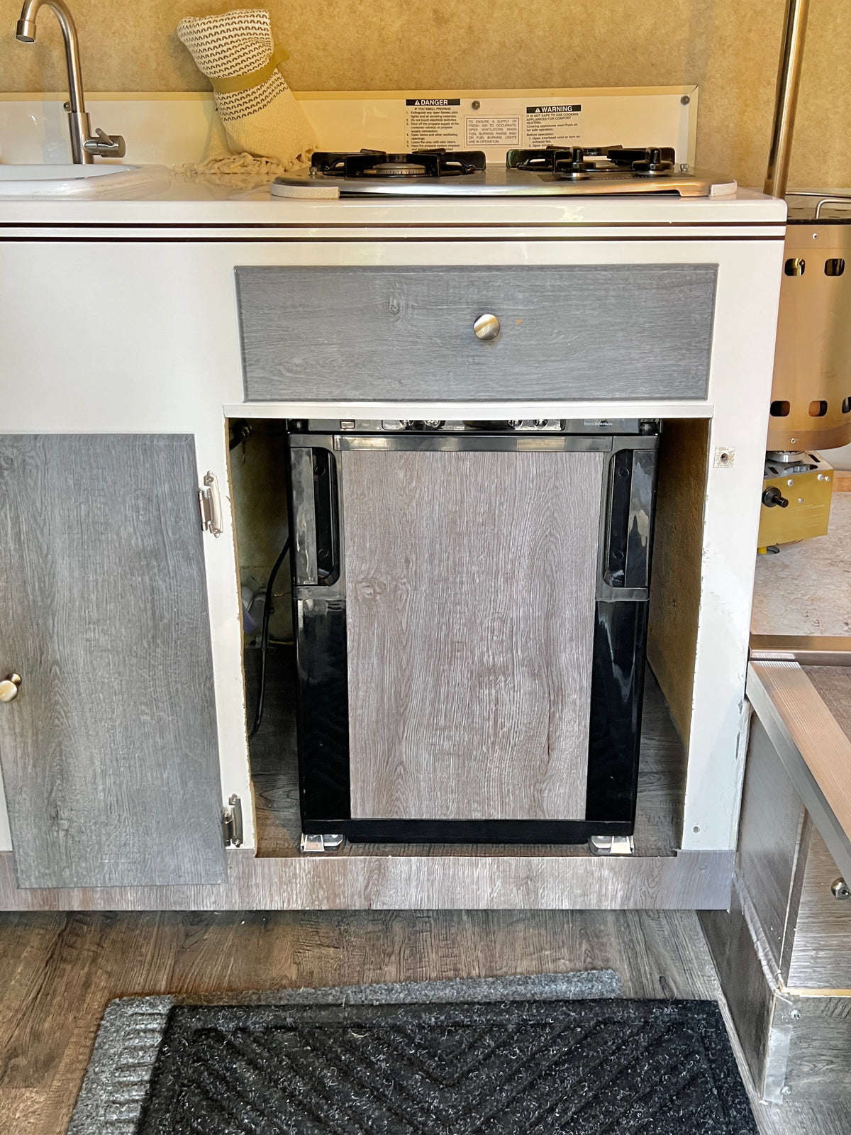 Compact RV rental kitchen features a white countertop with a sink, two-burner stove, and a wood-paneled mini-fridge below. — photo 9