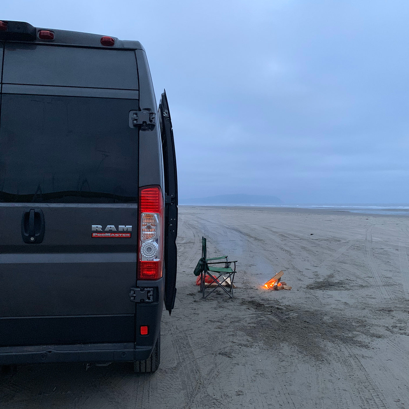 Dark grey RV rental van, a Ram ProMaster, parked on a vast sandy beach at dusk. A camping chair and glowing campfire are beside it.