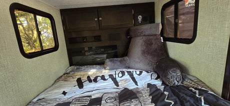 Cozy RV rental bedroom featuring a large bed with a backrest pillow, dark cabinetry, and bright windows showing lush trees outside.