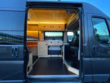 Open door view of a sleek grey RV rental's interior. Features include a custom wood bed, kitchen, Dometic fridge, and bright lighting.