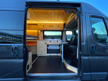 Open door view of a sleek grey RV rental's interior. Features include a custom wood bed, kitchen, Dometic fridge, and bright lighting.