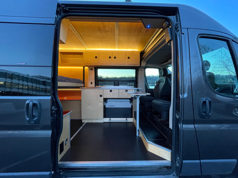 Open door view of a sleek grey RV rental's interior. Features include a custom wood bed, kitchen, Dometic fridge, and bright lighting. — photo 7