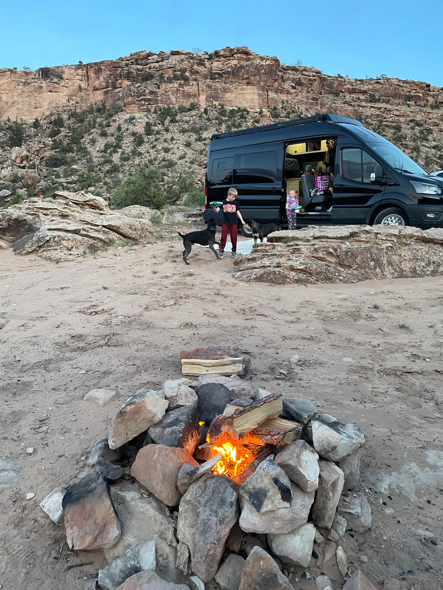 Enjoying an RV rental adventure: kids, dogs, and a campfire illuminate this desert campsite below rocky cliffs.