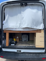 Inside an RV rental van: custom bed with privacy curtain, under-bed storage featuring a portable cooler.