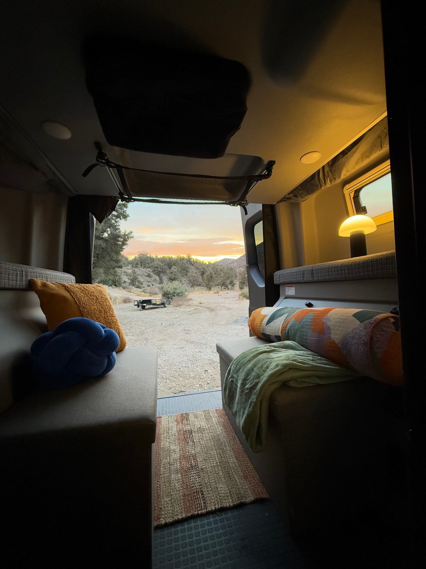 View from an RV rental: a cozy interior with pillows and a lamp, looking out at a serene campsite with a sunset sky. — photo 13