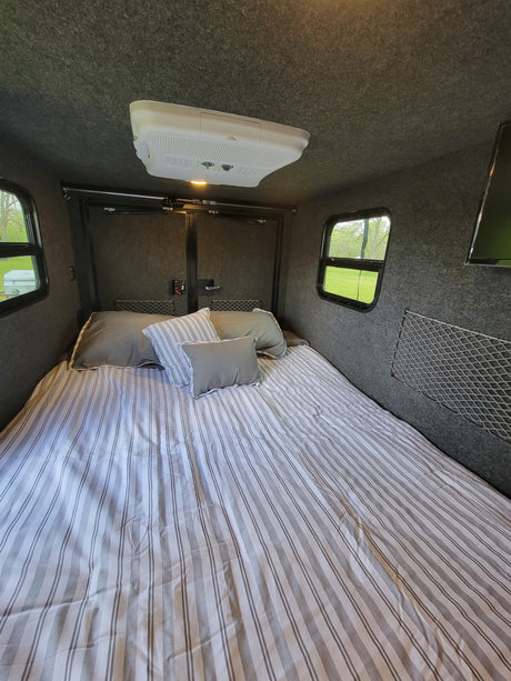 RV rental interior: cozy bedroom with gray striped bedding, carpeted walls, windows & ceiling AC.