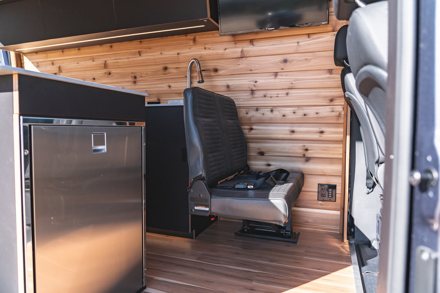 RV rental interior: Wood-paneled van with compact kitchen, stainless fridge, bench seat, and TV. Ready for adventure.
