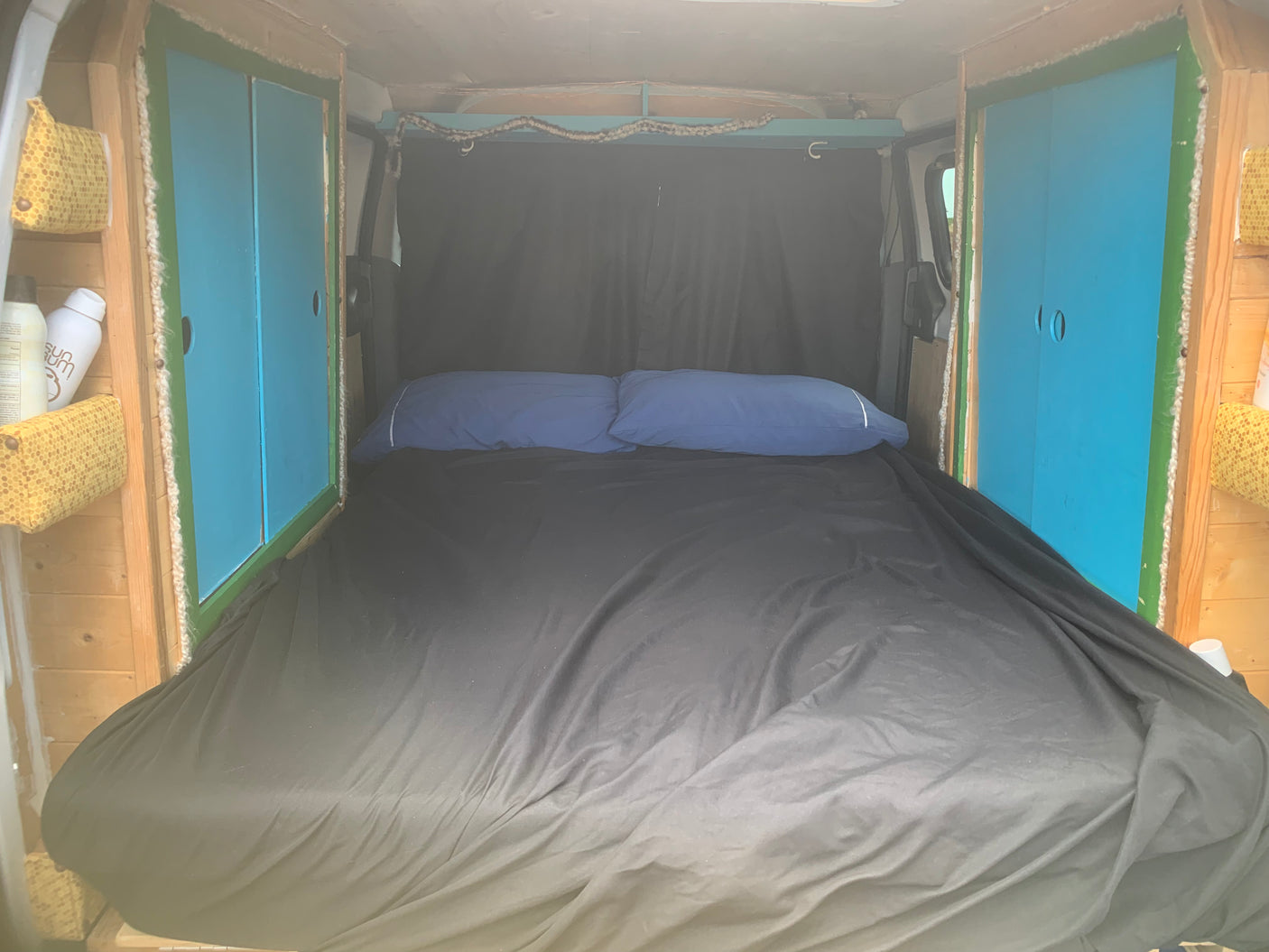 Inside a comfy RV rental camper, a full bed with black sheets & blue pillows. Wood panels & storage visible on sides.