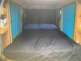 Inside a comfy RV rental camper, a full bed with black sheets & blue pillows. Wood panels & storage visible on sides.