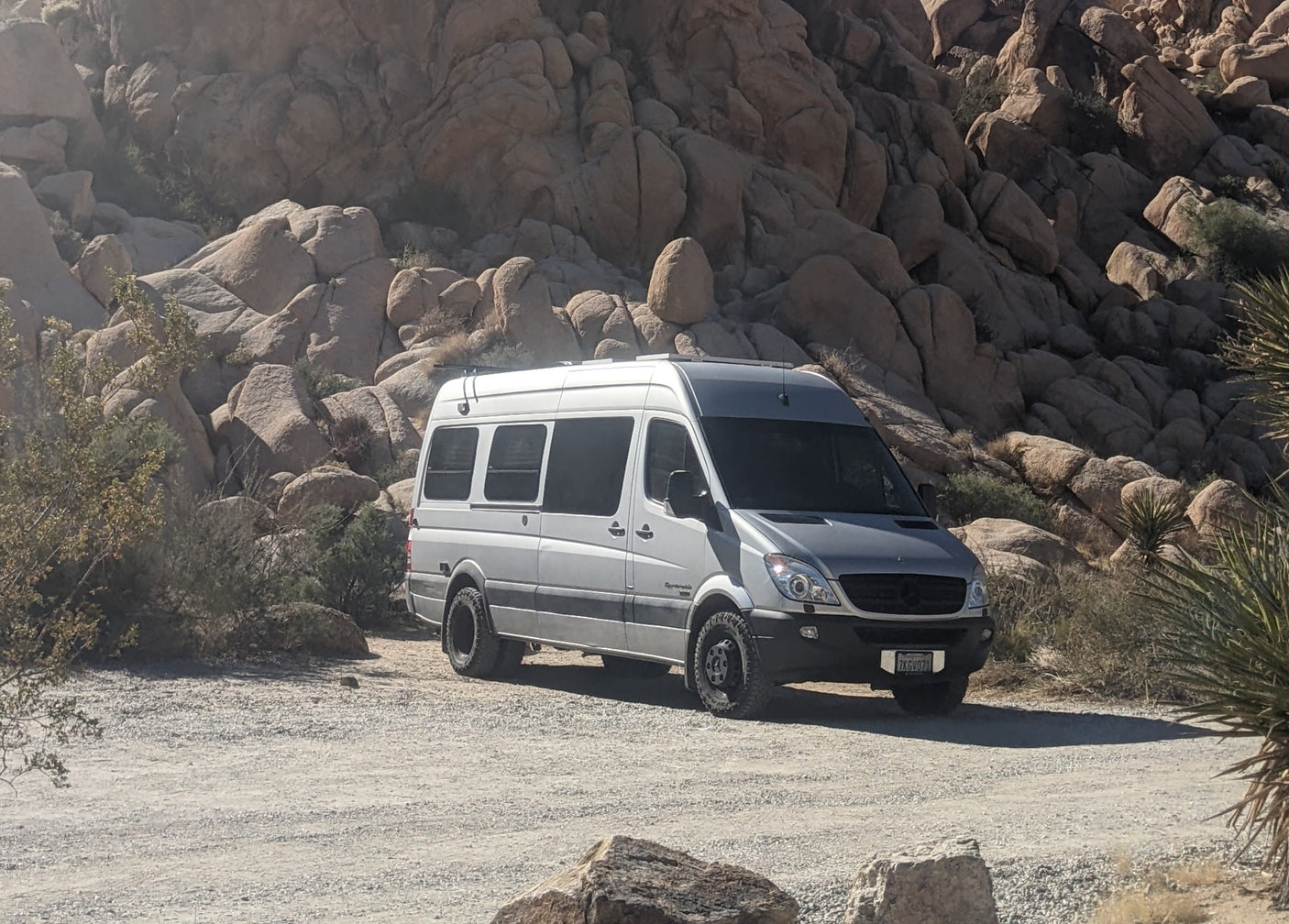 A silver Sprinter RV rental van on a dusty road, surrounded by large, rounded rock formations and desert plants.